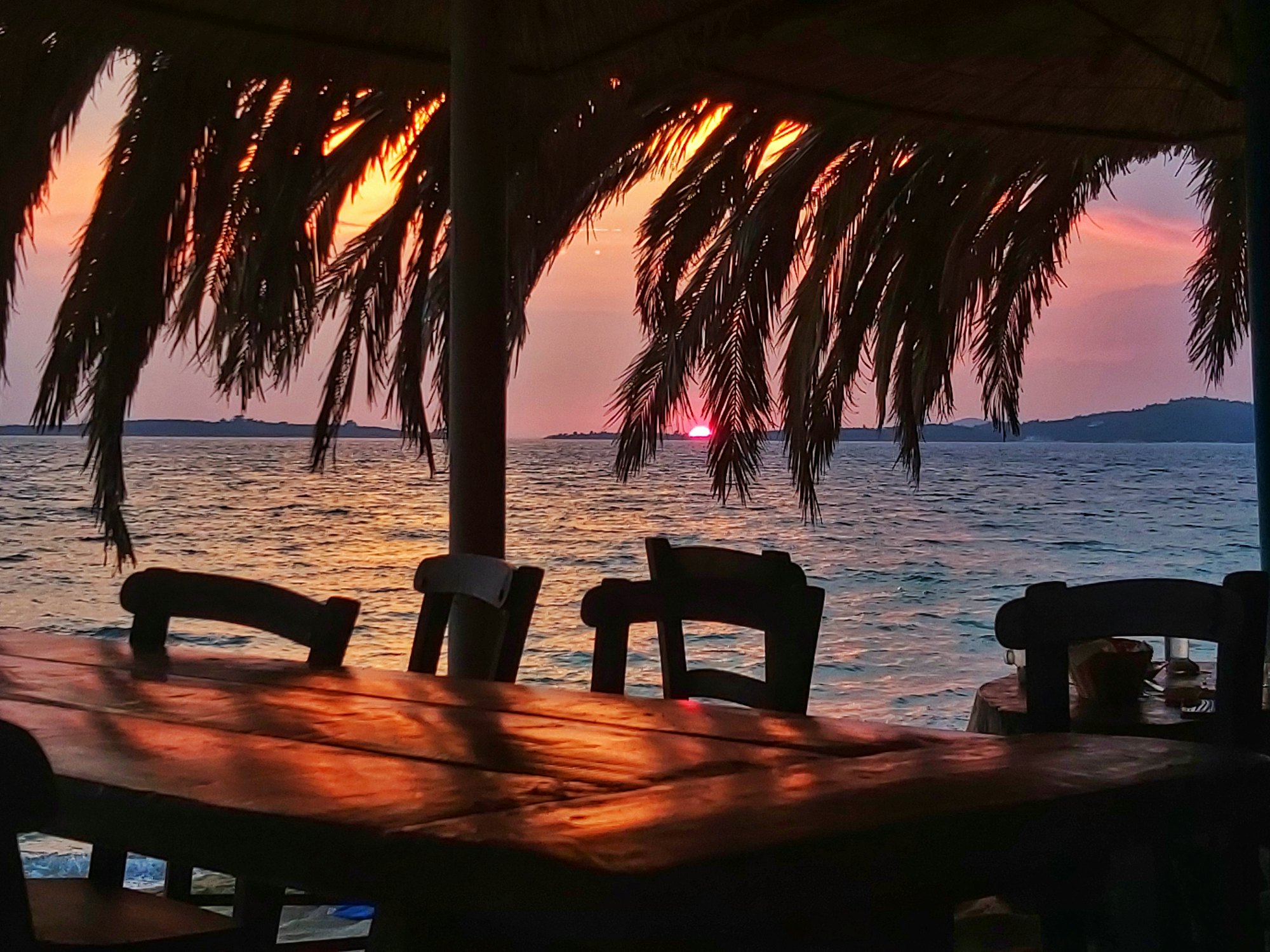 A thatched beach bar table looking out over the ocean as the sun sets.
