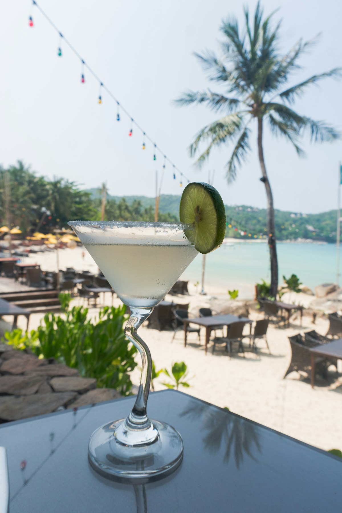 A cocktail on a table under a palm tree looking out at a tropical beach.